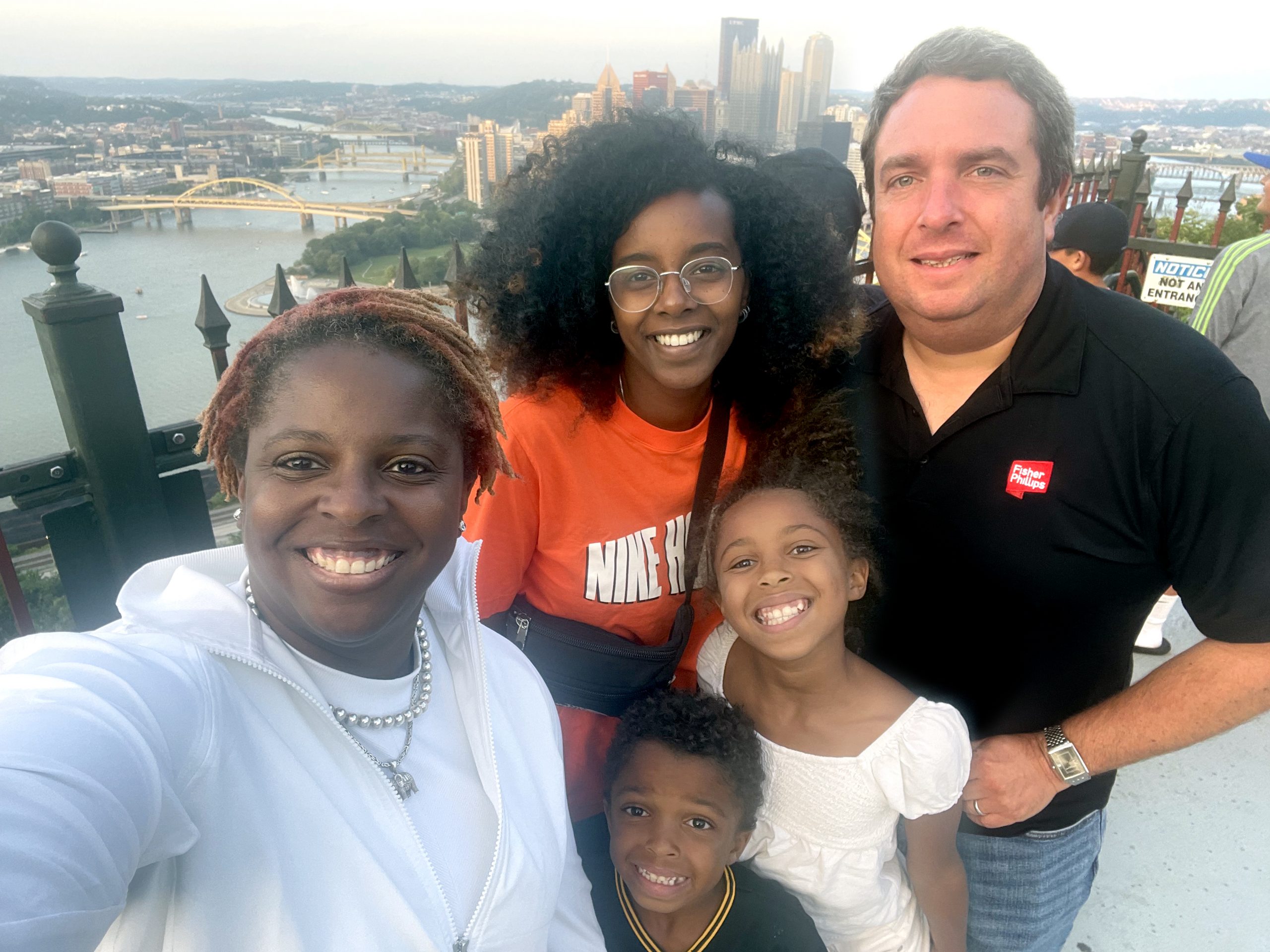 A selfie of a family, who is hosting a Shilchim, on top of Mount Washington with downtown Pittsburgh in the background.